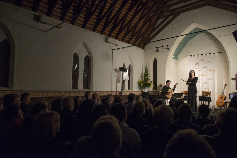 A woman and guitarist stand before a crowd in a church.